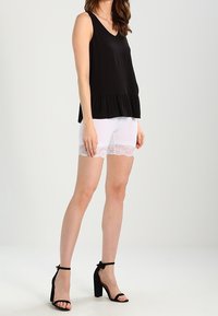 Femme portant un haut sans manches noir, un short blanc avec des bordures en dentelle, et des sandales à talons hauts noires, se tenant devant un fond uni.