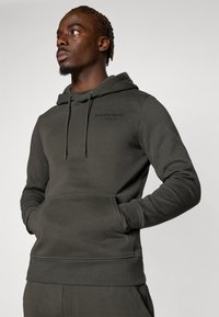 Donkergroene hoodie van zacht materiaal, voorzien van een kangoeroezak, een capuchon met trekkoord en een klein bedrukt logo op de borst.