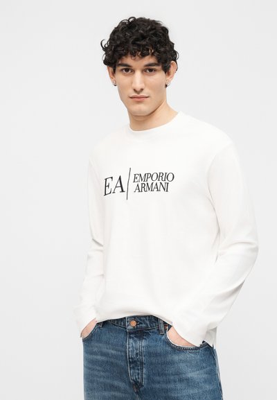 Νεαρός με σγουρά μαλλιά φορούντας ένα λευκό μακρυμάνικο πουκάμισο με το λογότυπο "EA Emporio Armani" και μπλε τζιν, με τα χέρια στις τσέπες, ουδέτερο φόντο.