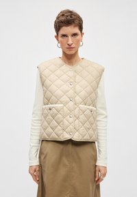 Colete bege acolchoado, design sem mangas, com botões de pressão e dois bolsos frontais. Usado sobre uma camisa branca de manga comprida.