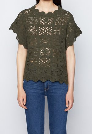 Oliwkowy top na szydełku z falbaniastym dekoltem i krótkimi rękawami z misternymi wzorami ażurowymi. W połączeniu z niebieskimi dżinsami.