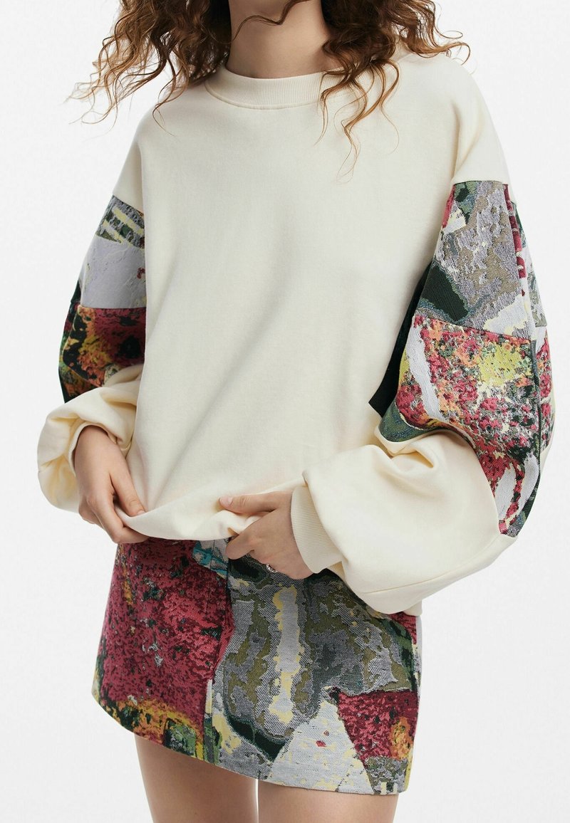 Femme portant un sweat crème avec des manches ballon à motifs abstraits colorés et une mini-jupe assortie à motifs.