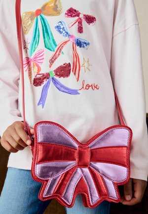 Borsa a forma di fiocco in velluto rosso e rosa con dettagli cuciti, tenuta da una persona che indossa una maglietta rosa chiaro con illustrazioni di fiocchi colorati.