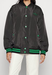 Svart satin bomberjacka med gröna tryckknappar och randiga ribbade ärmar och nederkant. Har broderad logotyp på bröstet.