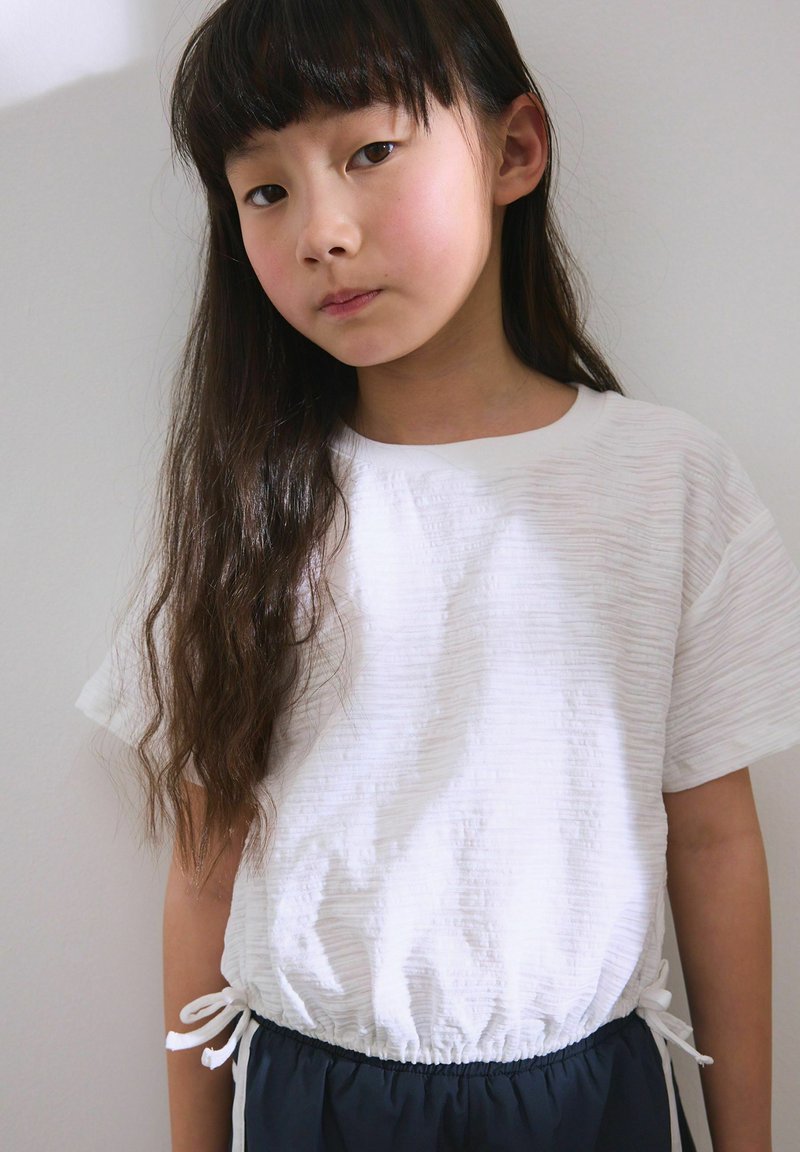 Jeune fille aux longs cheveux foncés portant un haut blanc texturé à manches courtes et un pantalon foncé, debout contre un mur clair uni.