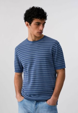 Giovane uomo con capelli neri ricci che indossa una maglietta a maniche corte a righe blu e bianche e jeans azzurri, con le mani nelle tasche, che guarda verso il basso.