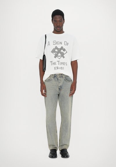 White graphic t-shirt with black print, paired with faded blue jeans. Black shoes and a small black bag complete the casual outfit.