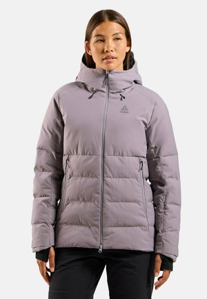 Frau trägt eine hellviolette Kapuzen-Winterjacke mit Reißverschlusstaschen und schwarze Hose, steht vor einem einfarbigen Hintergrund.