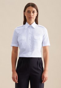 Camicia bianca a maniche corte con colletto a punta, due tasche sul petto e chiusura a bottoni distintiva, abbinata a pantaloni scuri plissettati.