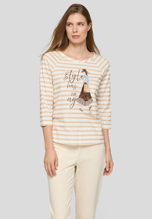 Frau trägt ein beige-weiß gestreiftes Shirt mit einer Illustration und dem Text „Stil kennt kein Alter“, kombiniert mit hellen Hosen.