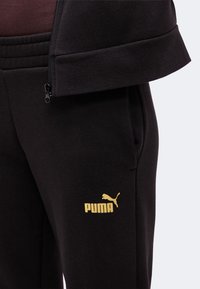 Μαύρη φόρμα Puma με χρυσλό λογότυπο Puma στον μηρό και μέρος μαύρου σακακιού με φερμουάρ ορατό πάνω από τη μέση.