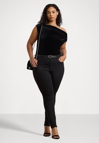 Svart sammetstopp med en axel och draperad design, tillsammans med svarta tighta jeans och en svart axelremsväska. Modellen har på sig svarta klackar med öppen tå.