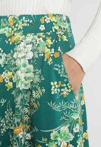 Una mano se adentra en el bolsillo de una falda color teal adornada con vibrantes patrones florales en tonos de verde, amarillo y blanco.