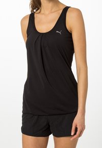 Camiseta atlética negra sin mangas con un diseño plisado, combinada con pantalones cortos de malla negra que presentan un patrón texturizado. Logo visible en la camiseta.