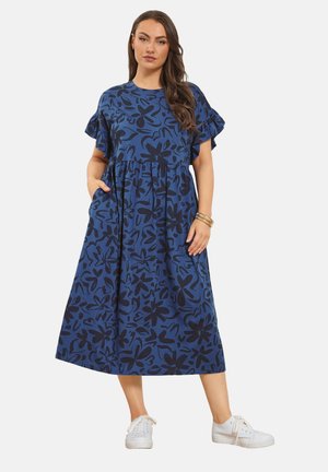 Kobieta ubrana w niebieską sukienkę midi z czarnym kwiatowym nadrukiem, z falbankami na rękawach, kieszeniami, białymi trampkami i złotymi bransoletkami, stojąca na białym tle.