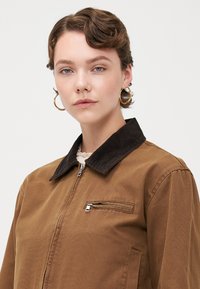 Jachetă zip-up maro cu un guler din material corduroy închis, având un buzunar pe piept și o textură moale. Modelul are părul scurt și cercei circulare.