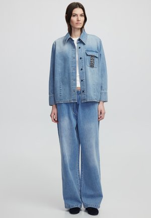 Veste en denim bleu clair avec un col pointu, fermeture à boutons sur le devant et une poche poitrine. Associée à un pantalon en denim large et décontracté.