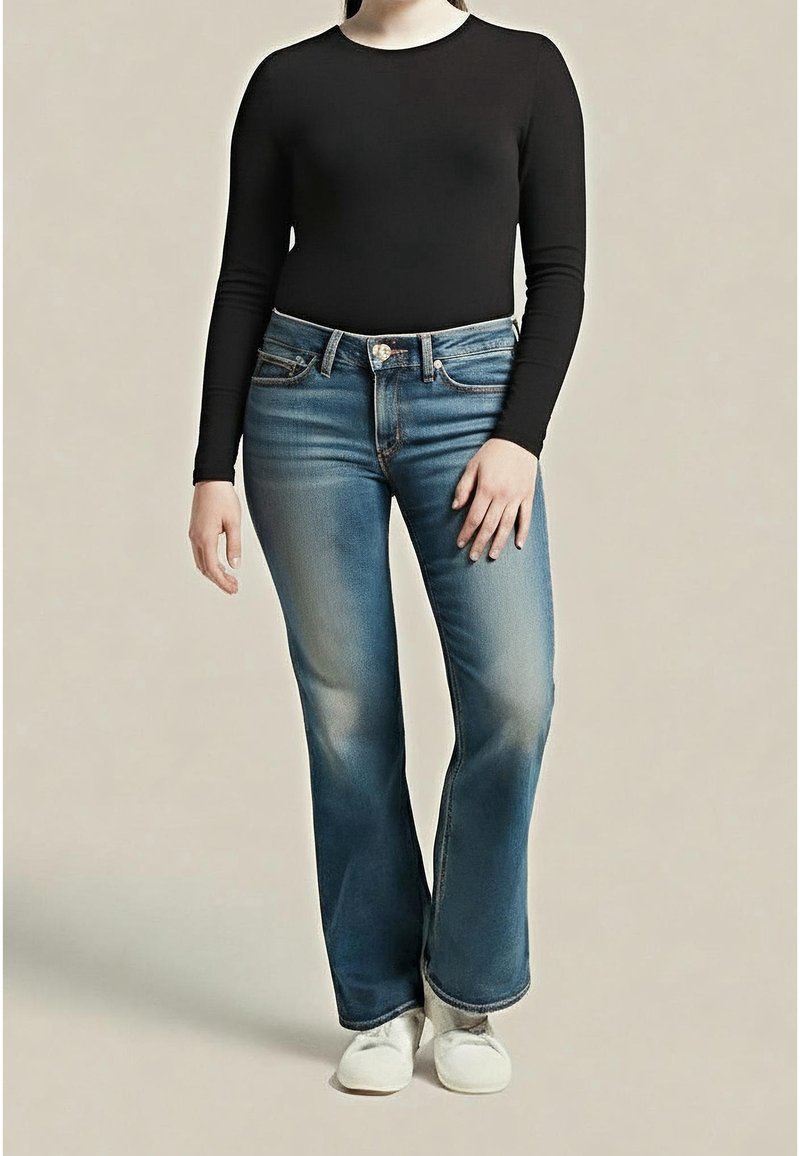 Personne portant un haut noir ajusté à manches longues, jean bootcut bleu délavé et chaussures blanches, debout devant un fond neutre.