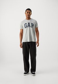 Graues Baumwoll-T-Shirt mit "GAP" in marineblauen Buchstaben, kombiniert mit schwarz weitem Jeans und schwarzen Sneakers. Modell trägt goldfarbene Sonnenbrille.