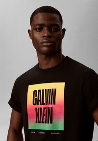 Jeune homme portant un t-shirt noir avec un grand carré multicolore et un texte en gras noir "CALVIN KLEIN" sur le devant.