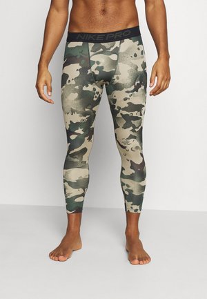 Männlicher Oberkörper und Beine, die grüne und beige camouflagegemusterte Nike Pro Kompressionsleggings tragen, barfuß auf einem hellgrauen Boden stehend.