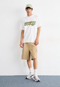Camiseta gráfica blanca con el logo "Carhartt WIP" en verde y amarillo, pantalones cortos tipo cargo en color beige, gorra verde claro y zapatillas con ribete plateado.