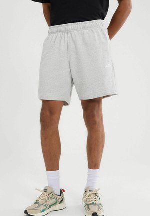 Mężczyzna ubrany w jasnoszare szorty z elastycznym pasem, białe skarpetki za kostkę oraz beżowe sportowe buty z zielonymi akcentami, stojący na jednolitym tle.