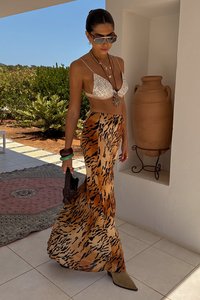 Top bikini con paillettes argentate, gonna a stampa animale in arancione e marrone, stivali con tacco beige e borsa nera. Ambiente esterno illuminato dal sole.