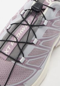 Helllila Sport Schuh mit Mesh-Obermaterial, strukturierten Überzügen, elastischen Schnürsenkeln und einer kontrastierenden weißen Zwischensohle. Zeigt Branding auf der Zunge.