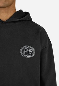 Čierna mikina s kapucňou vyrobená z jemného materiálu, s predným logom "PEGADOR" a textom "SNEŽIENKY PADÁVAJÚ, DOBRODRUŽSTVÁ RASTÚ" v bielej farbe.