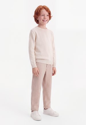 Barn med rødt hår smiler iført en lysebeige sweater, matchende beige bukser og hvide sneakers, stående mod en hvid baggrund.