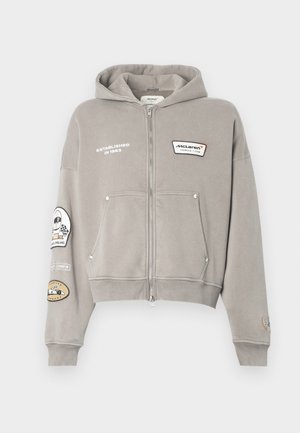 Gray zip-up hoodie in soft fabric, featuring a hood, two front pockets, and various embroidered patches on sleeves and chest.
