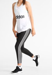 Camiseta sin mangas blanca con el logo negro de "adidas", combinada con mallas negras que tienen tres franjas blancas, y zapatillas deportivas negras.