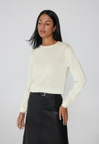 Suéter de manga larga color crema claro con un cuello redondo, hecho de una tela suave y texturizada, combinado con una falda de cuero negra con cinturón.