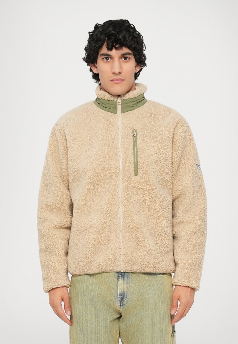 Beige fleeced jack met een hoge kraag, groene ritsaccenten en een borstzak aan de voorkant. Zachte textuur en een relaxte pasvorm met lange mouwen.