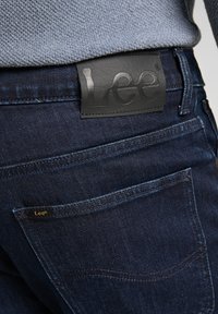 Mörkblå denimjeans med en slät textur, som har en svart läderlogotyp på midjebandet och subtila sömnadsdetaljer.