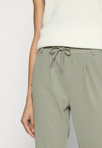 Calças verticais de cor verde-claro, com cintura ajustável por cordão e detalhes prateados. Apresentadas com um top de malha branco, o foco está na textura das calças.