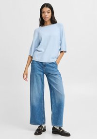 Svetle modrý pletený svetr s volným střihem a širokými rukávy, v kombinaci s džíny s vysokým pasem a širokými nohavicemi v modré barvě a tmavými mokasíny.