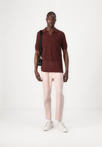 Polo bourgogne à manches courtes avec un motif texturé, associé à un pantalon tapered rose clair et des baskets blanches, se tenant contre un fond neutre.