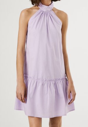 Femme portant une robe légère violette sans manches avec un col halter froncé et un ourlet à volants, debout devant un fond uni.