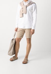 Camicia bianca con bottoni, pantaloni corti beige, maglione beige posato sulle spalle, che porta una borsa tote beige chiaro e indossa sandali color beige.
