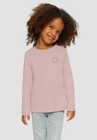 Jong kind met krullend haar, gekleed in een lichtroze shirt met lange mouwen en lichtblauwe jeans, glimlachend en iets opzij kijkend.
