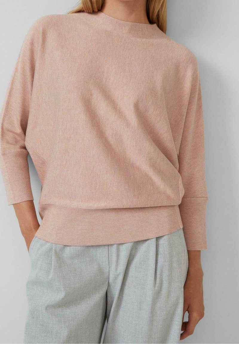 Femme portant un pull en tricot rose clair à manches trois-quarts et un pantalon plissé gris clair, les mains dans les poches.