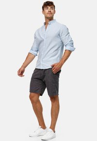 Lichtblauw en wit gestreept overhemd met opgerolde mouwen, gecombineerd met donkergrijze shorts en witte sneakers. Casual zomeroutfit.