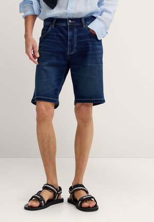 Mann trägt dunkelblaue Jeansshorts, hellblaues langärmeliges Hemd, das in die Hose gesteckt ist, und schwarze Sandalen mit verstellbaren Riemen.