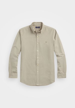 Chemise beige à manches longues avec col boutonné, arborant un petit logo sur la poitrine gauche et en tissu coton doux.