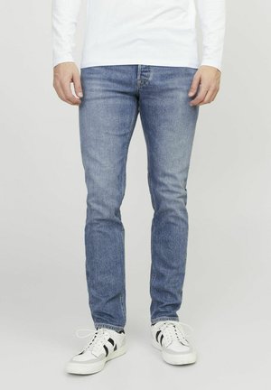 Jeans de denim azul claro con un corte ajustado, que presentan un desgaste sutil y detalles de costura. Combinados con zapatillas blancas y una camiseta de manga larga blanca.