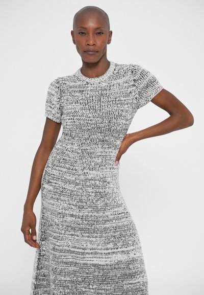 Donna con testa rasata che indossa un vestito a maglia bianco e nero a maniche corte, in posa con una mano sui fianchi su uno sfondo bianco semplice.