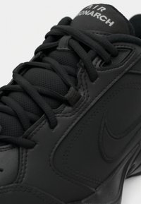 Close-up do ténis Nike Air Monarch preto, mostrando os atacadores, língua texturizada e o logótipo Nike swoosh bordado no painel lateral.
