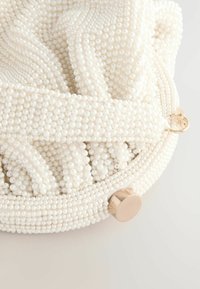 Valkoinen helmillä varustettu clutch-laukku, jossa on teksturoitu pinta pienistä pyöreistä helmistä ja metallinen suljenta. Laukku on pyöreä muodoltaan ja siinä on rypistetty kangas.